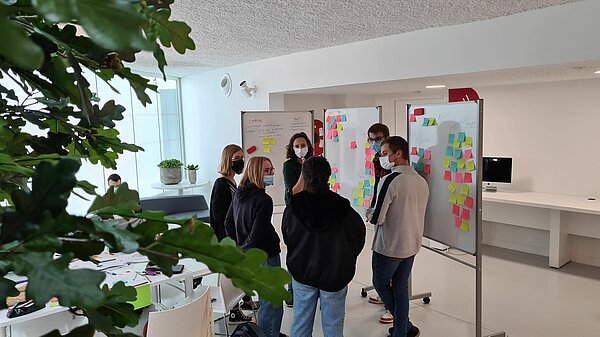 Un groupe de six personnes dans le salon P.I., devant des tableaux blancs remplis de post-it, discutant ensemble pour préparer une présentation