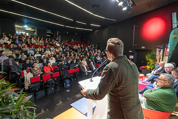 Amphi A du pôle événementiel lors d'une conférence, photo prise depuis la scène pour montrer le public avec le conférencier de dos 