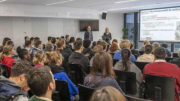 Une présentation réalisée par deux personnes dans l'amphi B du pôle événementiel, avec le public de dos