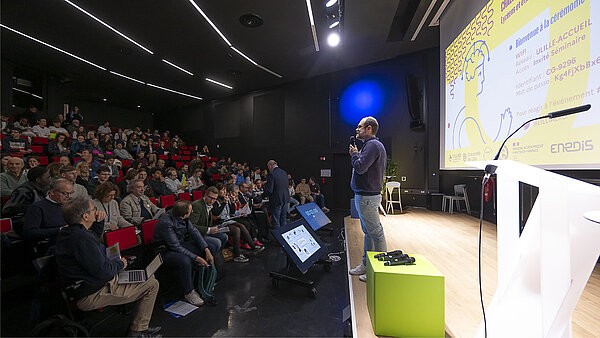 Une vue latérale de la scène de l'amphi A du pôle événementiel, avec le présentateur sur scène à droite de l'image et le public assis à gauche de l'image 