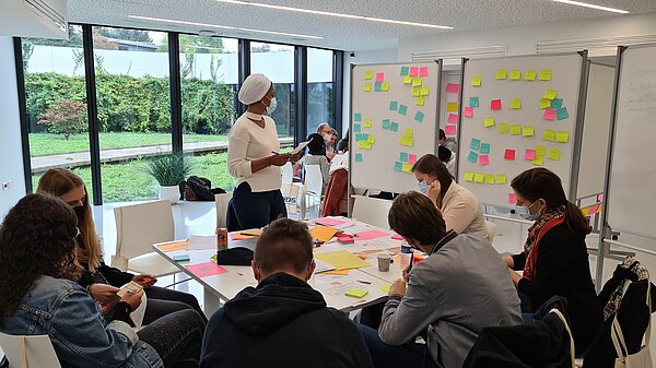 Un groupe de travail autour d'une table dans la salle D, une personne est debout devant deux tableaux blancs remplis de post-it