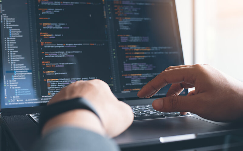 Des mains en premier plan tapent sur le clavier d'un ordinateur portable. L'écran de l'ordinateur montre un logiciel de code informatique.
