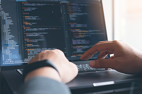 Des mains en premier plan tapent sur le clavier d'un ordinateur portable. L'écran de l'ordinateur montre un logiciel de code informatique.